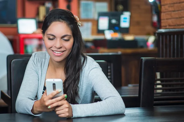 Aantrekkelijke brunette dragen grijze trui zitten door restaurant tabel kijken naar mobiele telefoon — Stockfoto