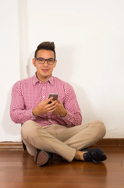 Hispanic male wearing red white shirt, khaki jeans sitting on wooden floor against wall pressing mobile phone, empty background — Stock Photo, Image