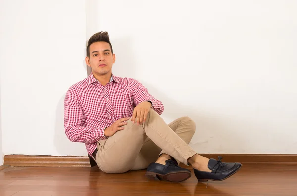 Homem hispânico vestindo camisa branca vermelha sentado no chão de madeira contra parede fundo vazio — Fotografia de Stock