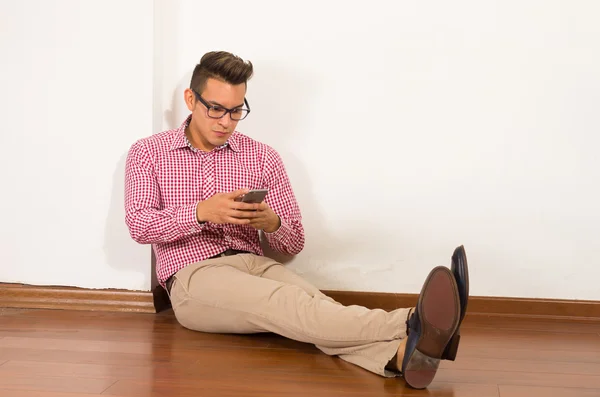 Spanischer Mann mit rotweißem Hemd, khakifarbener Jeans sitzt auf Holzboden gegen Wand drückendes Mobiltelefon, leerer Hintergrund — Stockfoto