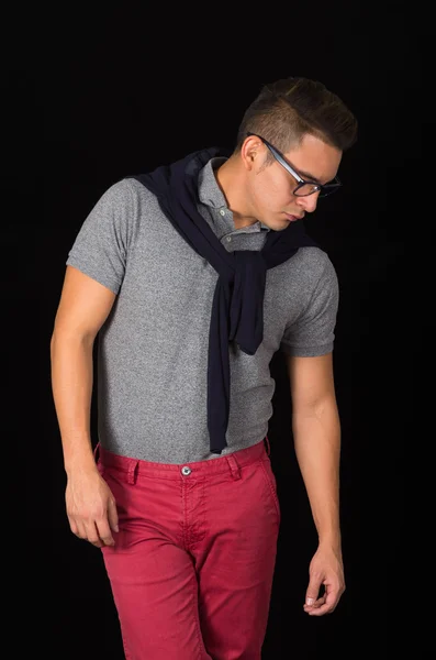 Hombre hispano con camisa ajustada, suéter sobre los hombros, pantalones rojos y gafas que retratan un estilo hipster sofisticado, mirando hacia abajo, fondo negro —  Fotos de Stock