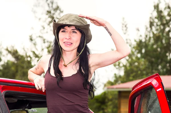 Morena aventurera con sombrero de safari verde, ambiente al aire libre de pie en la puerta roja del coche mirando hacia adelante explorando —  Fotos de Stock