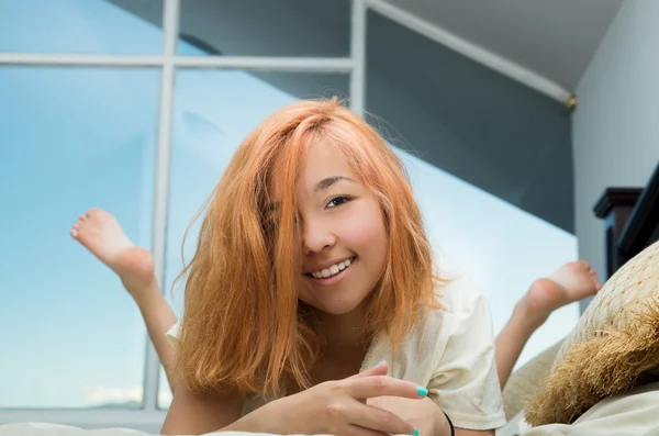 Pretty young woman lying comfortably on bed smiling to camera with large windows in background — ストック写真