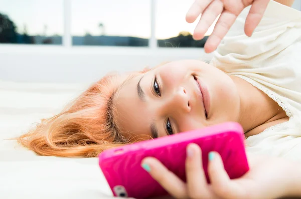 Jolie jeune femme couchée confortablement sur le lit et utilisant un téléphone portable rose avec de grandes fenêtres arrière-plan — Photo