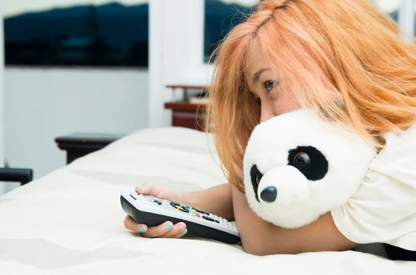 Mujer bastante joven acostada cómodamente en la cama abrazando al animal panda relleno y presionando el control remoto —  Fotos de Stock