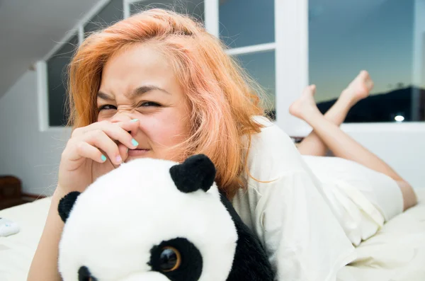 Pretty young woman lying comfortably on white bed and hugging stuffed panda animal with large windows in background — ストック写真