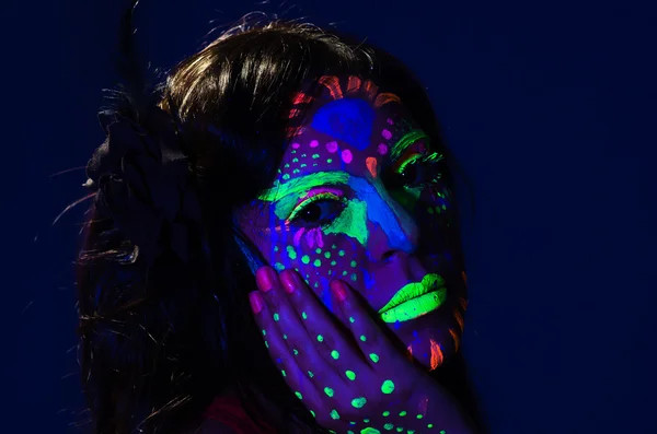 Headshot woman wearing awesome glow in dark facial paint, blue based with other neon colors and obscure abstract background, facing camera — Stock Photo, Image