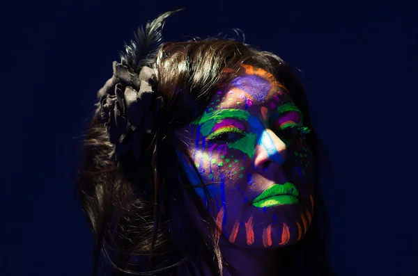 Headshot woman wearing awesome glow in dark facial paint, blue based with other neon colors and obscure abstract background, facing camera — Stock Photo, Image