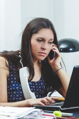 Hispanic brunette sitting by office desk talking on telephone with occupied and worried facial expression