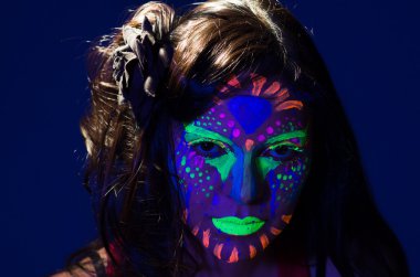 Headshot woman wearing awesome glow in dark facial paint, blue based with other neon colors and obscure abstract background, facing camera