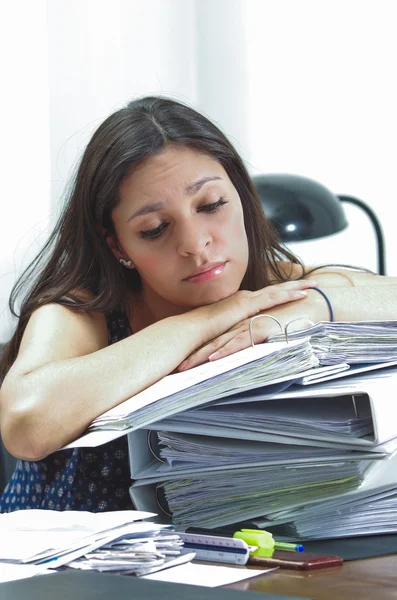 Hispanic brunette office worker sitting by desk and lying over pile of archive files looking tired — ストック写真