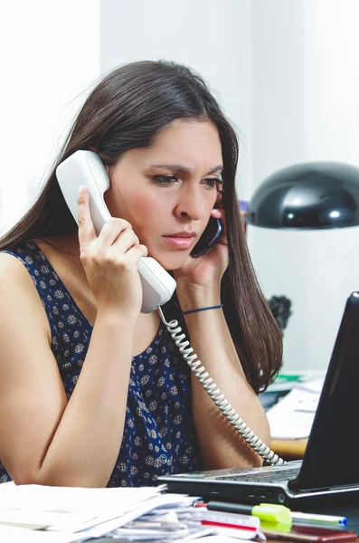 Morena hispânica sentada na mesa do escritório conversando por telefone com expressão facial ocupada e preocupada — Fotografia de Stock