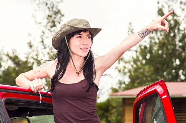 Adventurous brunette wearing green safari hat, outdoors environment standing in red car door looking forward scouting — Stok fotoğraf
