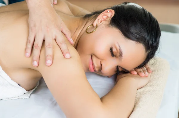 Hispanic brunette model getting massage spa treatment, white towel covering upper body lying horizontal smiling to camera with hands working on shoulders — 图库照片