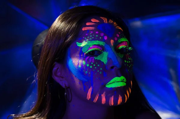 Headshot woman wearing awesome glow in dark facial paint, blue based with other neon colors and obscure abstract background, facing camera — Stockfoto