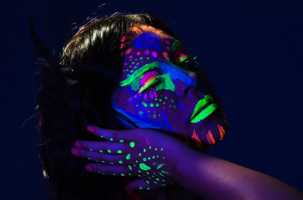 Headshot woman wearing awesome glow in dark facial paint, blue based with other neon colors and obscure abstract background, facing camera — Stock Photo, Image