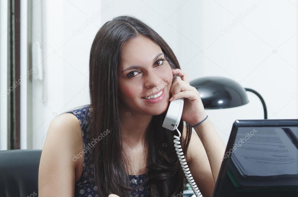 Hispanic brunette sitting by office desk talking on telephone smiling with positive attitude