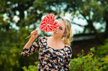 Bahçe ortamında, mutlu bir şekilde kamera için poz büyük lolipop tutan yazlık elbise giyen güzel model