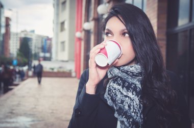 Zarif kadın kupa kahve içme kentsel ortamda koyu ceket ve beyaz siyah eşarp giyiyor