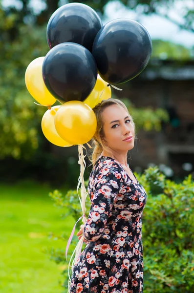 Beautiful hispanic model wearing summer dress in garden environment holding up golden and black balloons while posing for camera — Stock Photo, Image