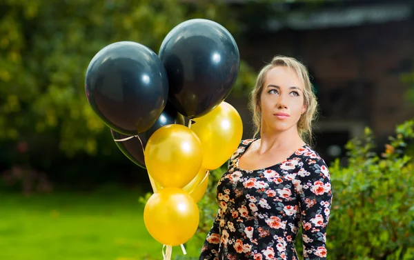 Vackra spansktalande modell iklädd sommarklänning i trädgårdar miljö håller upp golden och svarta ballonger medan poserar för kameran — Stockfoto