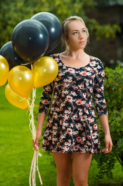 Krásný hispánský model nosí letní šaty v prostředí zahrady zvedl zlaté a černé balónky při pózování pro fotoaparát — Stock fotografie