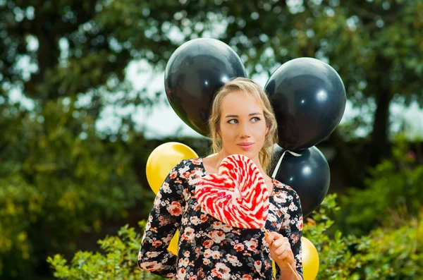 Vacker modell iklädd sommarklänning i trädgårdar miljö, svart gyllene ballonger och stora lollipop glatt poserar för kameran — Stockfoto