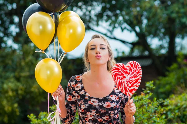 Modelo bonito vestindo vestido de verão no ambiente do jardim, segurando balões dourados pretos e grande pirulito posando feliz para a câmera — Fotografia de Stock