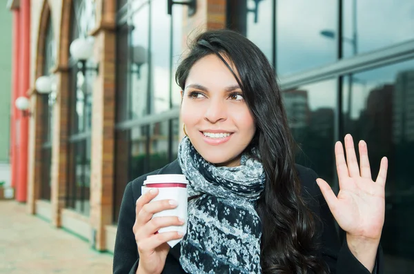 Donna bruna di classe che indossa un cappotto scuro e una sciarpa nera bianca in ambiente urbano con una tazza di caffè in posa per la fotocamera — Foto Stock