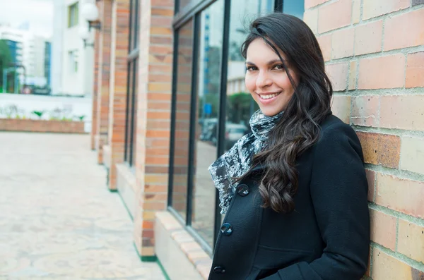 Classy woman wearing dark coat and black white clothing urban environment standing with back against brick wall smiling to camera — Stock Photo, Image