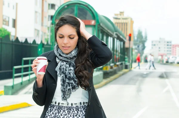 Mulher elegante vestindo casaco escuro e preto branco ambiente urbano roupas segurando caneca de café, posando com expressão facial decepcionada na frente da estação de ônibus — Fotografia de Stock