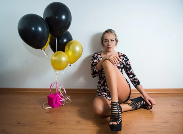 Vacker modell sitter på trägolv lutar sig tillbaka mot vit vägg klädd sommarklänning, höga klackar, svarta och gyllene ballonger hängande i luften knutna till rosa nuvarande box — Stockfoto