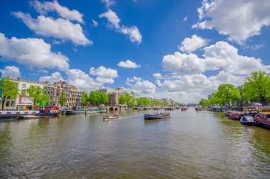 Amsterdam, Netherlands - July 10, 2015: Large water channel running through city with several boats parked alongside, green trees sourrounding and beautiful blue sky clipart