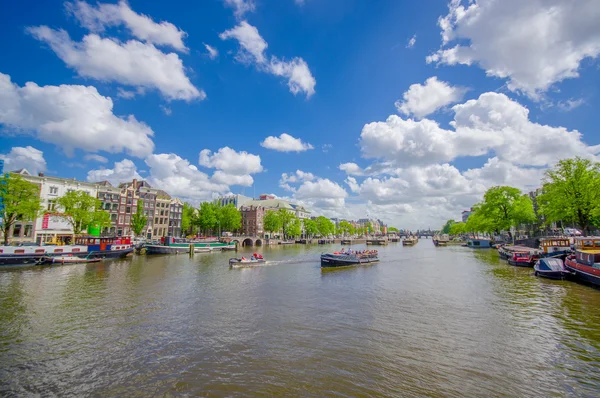 Amszterdam, Hollandia - 2015. július 10.: Nagy víz-csatorna fut keresztül a város, gyönyörű kék ég és zöld fák (betûi) melletti leparkolását több hajó — Stock Fotó