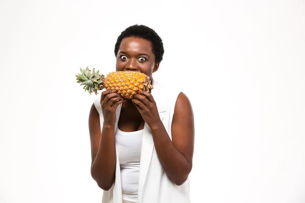 Hungry excited african american woman holding and biting fresh pineapple — стокове фото