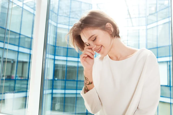 Mujer sonriente de pie cerca de la ventana en la oficina y riendo —  Fotos de Stock