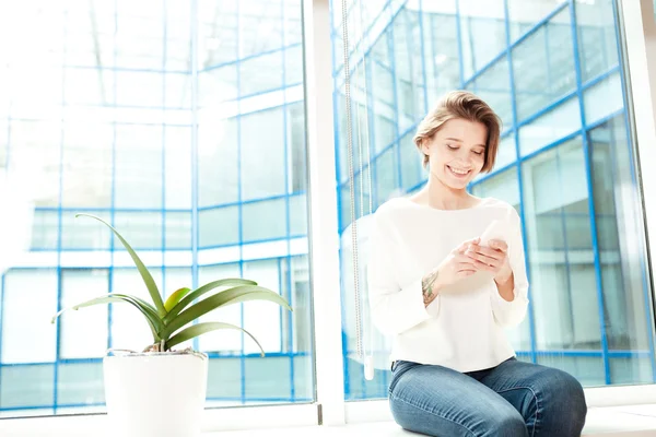 Mujer alegre sentado en el alféizar de la ventana en la oficina y el uso de teléfono inteligente —  Fotos de Stock