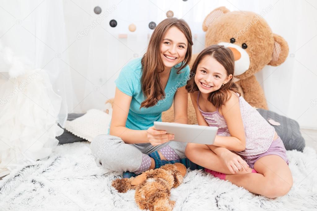 Two charming smiling sisters sitting and using tablet together 