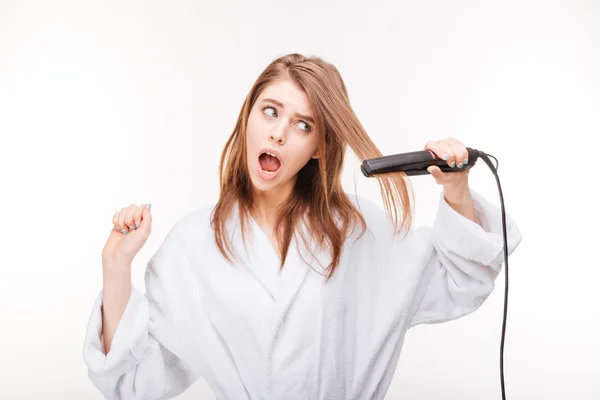 Налякана красива молода жінка в халаті за допомогою випрямлення волосся — стокове фото