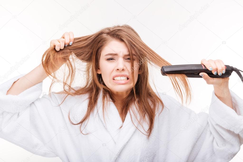 Angry irritated young woman straightening her hair using straightener 