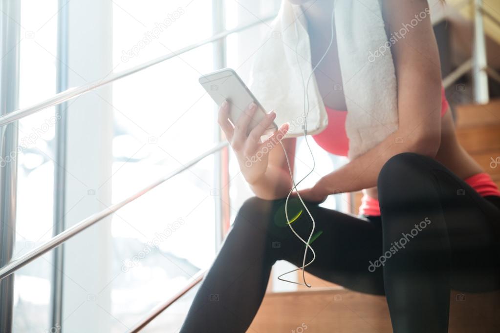 Sportswoman sitting and using smartphone with earphones in gym