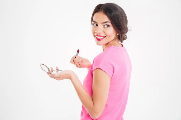 Cheerful pretty young woman holding mirror and applying lipstick — Stock Photo, Image