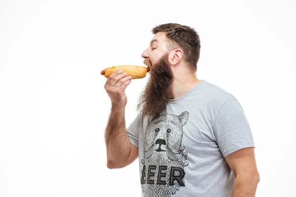 Bonito homem com barba de pé e comer cachorro quente — Fotografia de Stock