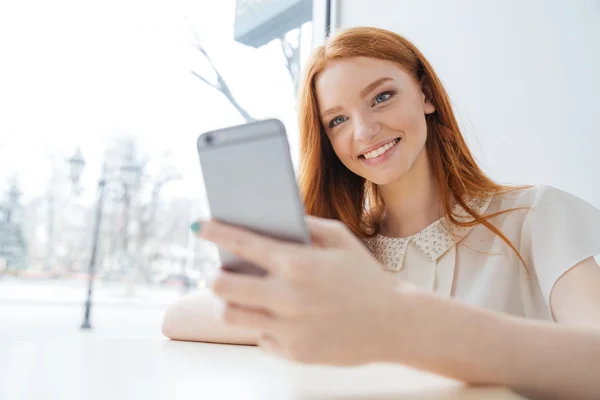 Веселая женщина с длинными рыжими волосами с помощью смартфона в кафе — стоковое фото