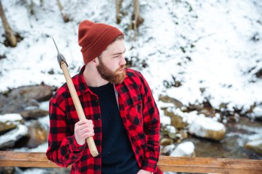 Pensive man in checkered shirt holding axe at winter forest clipart
