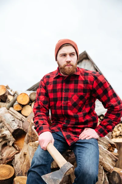 Ernster junger gutaussehender Mann sitzt auf Baumstämmen und hält Axt — Stockfoto