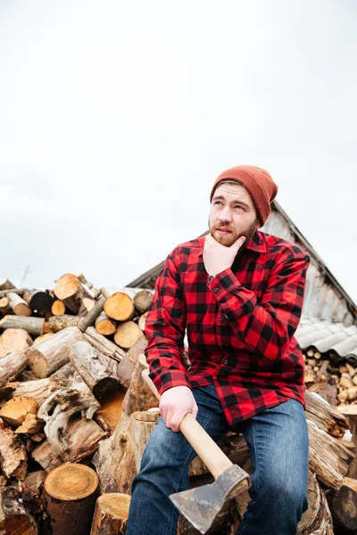 Nachdenklicher bärtiger Mann mit Axt sitzt auf Baumstämmen und denkt nach — Stockfoto