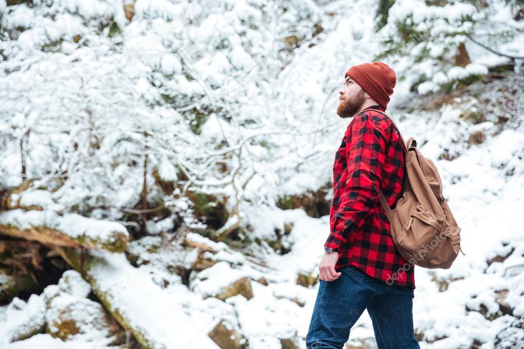 Serious bearded young man with backpack walking in winter forest