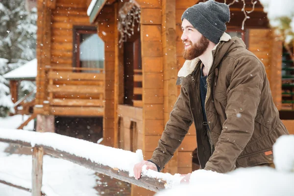 Lächelnder bärtiger Mann steht im Winter in der Nähe von Holzhäuschen — Stockfoto