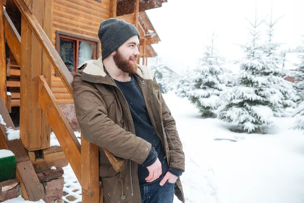 Nachdenklicher Mann mit Bart steht im Winter neben Blockhaus — Stockfoto
